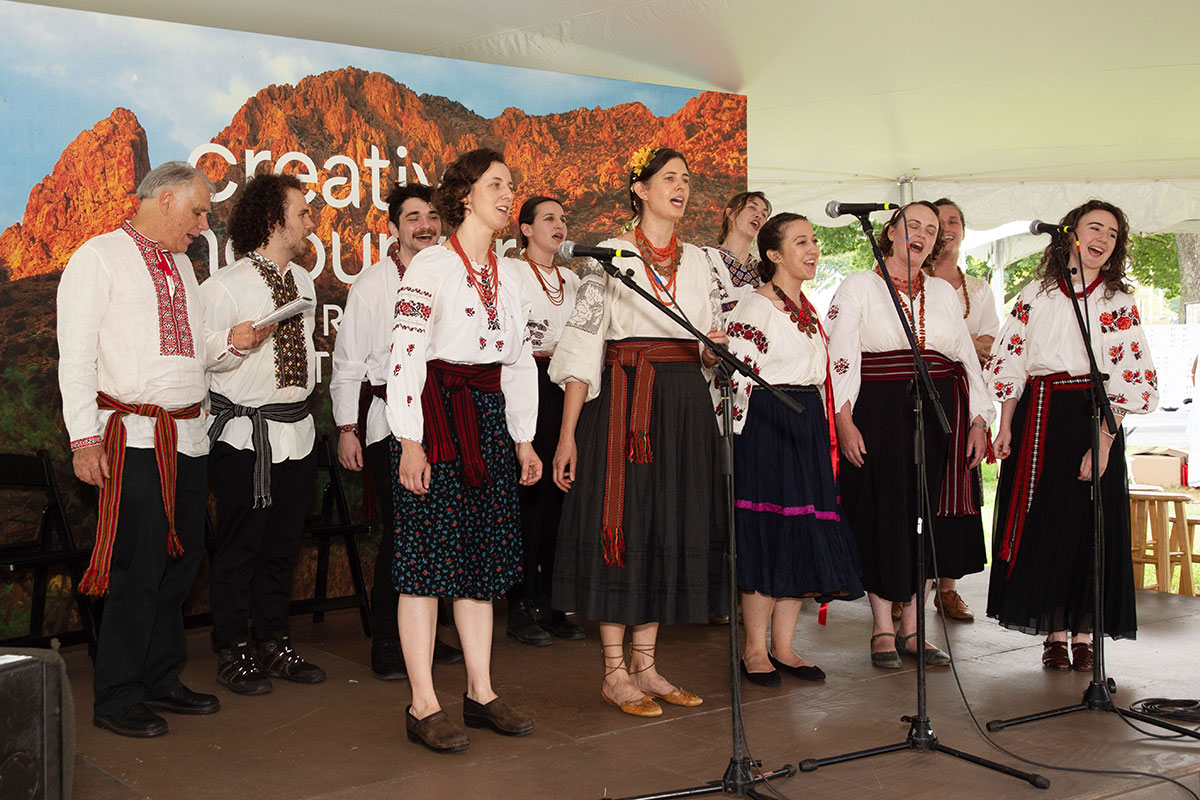 Eleven people sing on an outdoor stage, all wearing white embroidered shirts and dark skirts or pants.