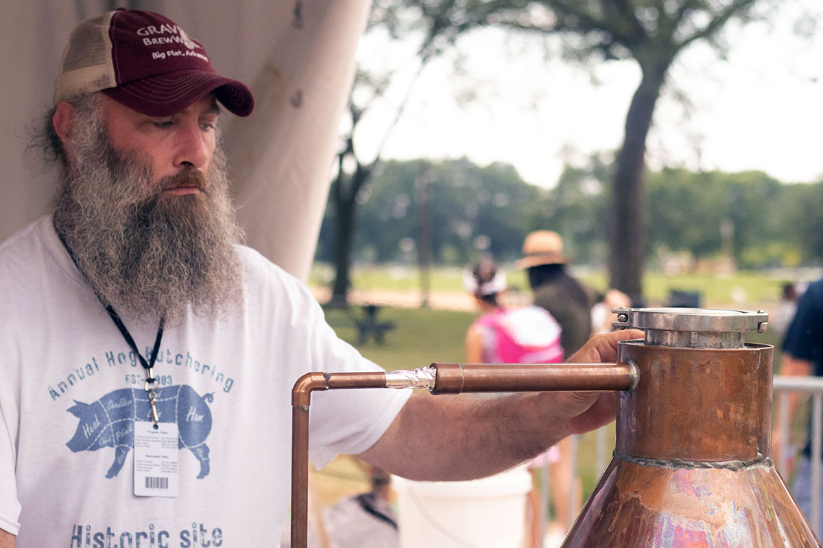 A man with a big gray beard and Annual  Hog Butchering T-shirt inspects a copper still outdoors.
