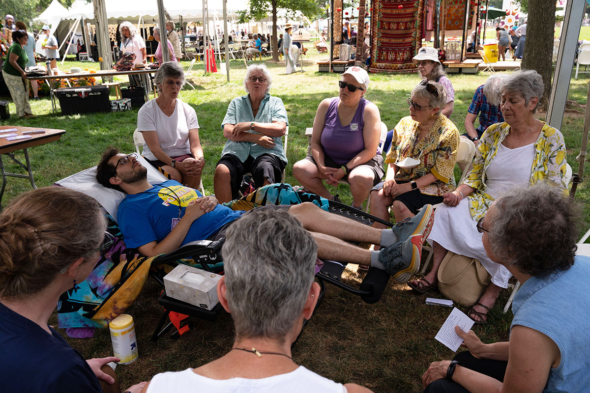 A young man in a Folklife Festival staff T-shirt reclines in a chair, eyes closed, as the people seated around him sing. They are under a tent in the grass, with other festival tents in the background. 