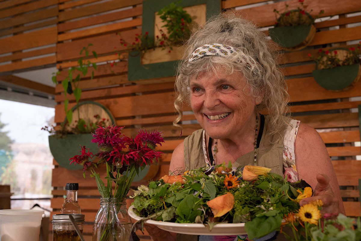 A woman with wavy gray hair smiles, holding up a white dish piled high with salad greens and yellow and orange edible flowers.