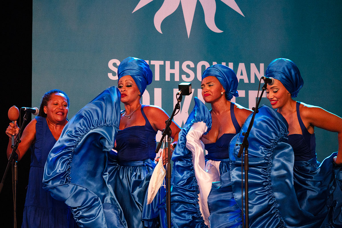 Three women in matching brilliant blue flared dresses dance on stage, next to another woman playing maracas.
