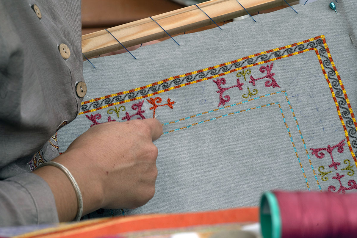 Closeup of someone embroidering a colorful border on pale blue fabric sewn to a wooden frame.