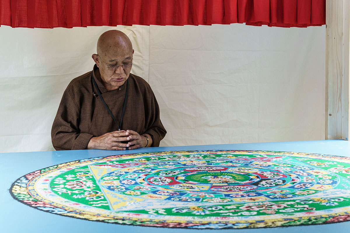 A man in a brown shirt stands before a circular mandala on a table, with his head bowed and hands together.