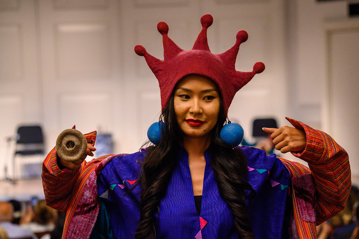 A young Asian person with long dark hair walks in a fashion show, wearing a bright pink, five-pointed felt hat, embroidered blue shirt and striped red jacket, oversize blue felt ball earrings, and an oversize gray circular ring. 