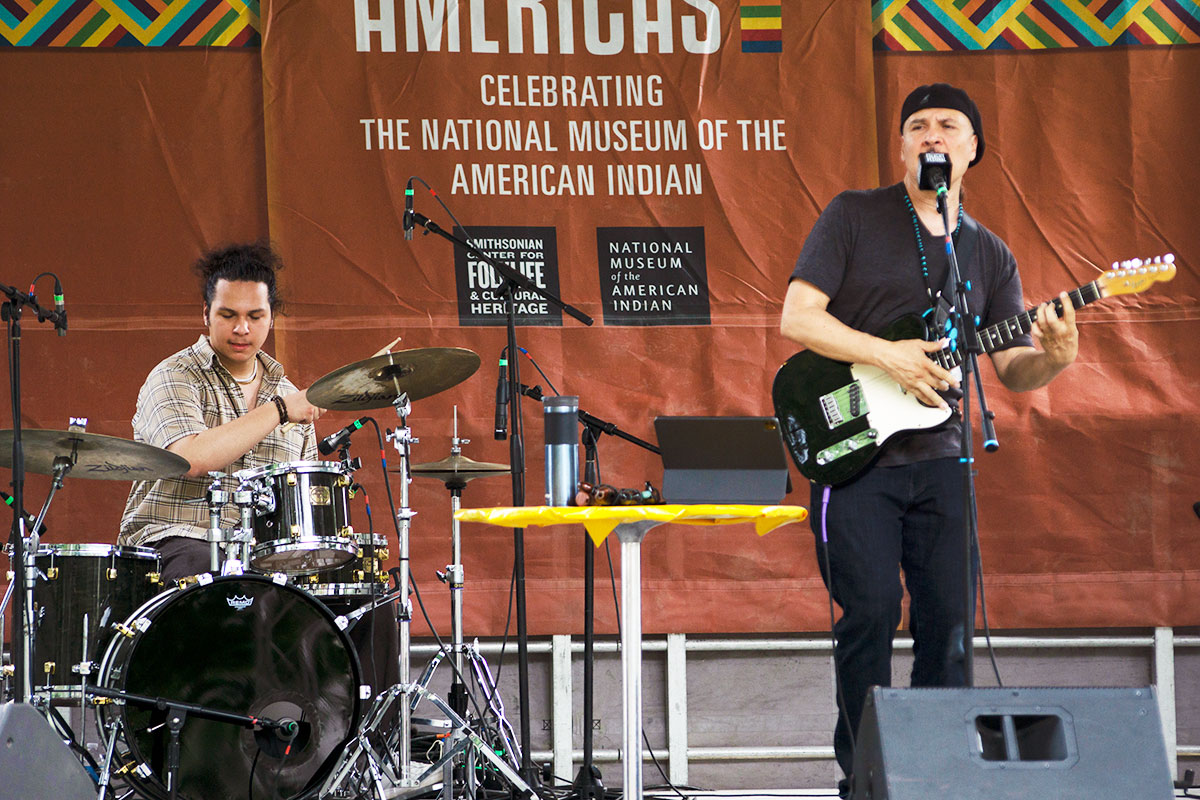 On an outdoor stage with an orange backdrop, a young man plays a drum kit while an older man, dressed in all black, playing a black electric guitar and sings into a microphone.