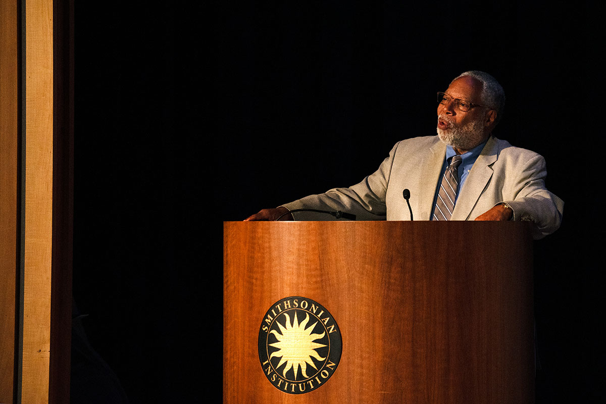 A man in a gray suit speaks on a stage at a podium with the Smithsonian sun logo in gold and black.