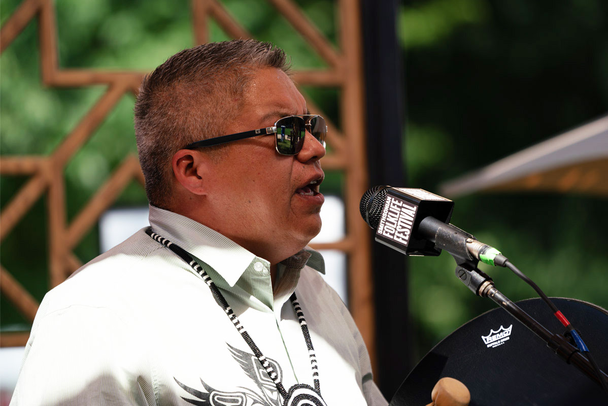 A man wearing a white shirt and sunglasses sings into a microphone