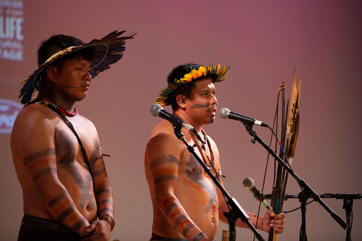 Two men stand on stage in front of microphone. Both wear black and red body paint on their bare chests and faces and visor-like crowns made of bird feather. One holds a wooden bow and arrows upright in his hand.