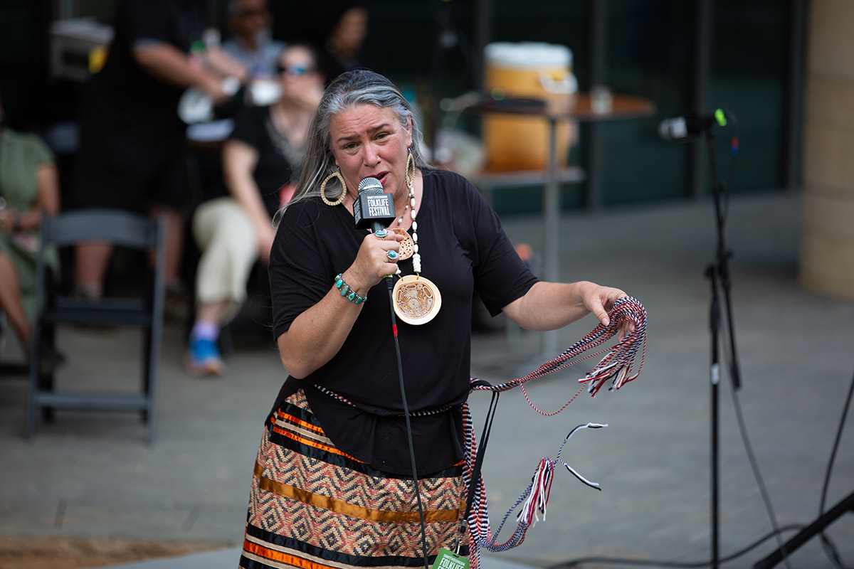 A woman with long, gray hair wearing a black top, orange patterned skirt, and a beaded necklace stands holding a microphone.