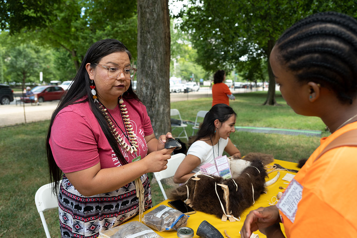 A woman wearing a red T-shirt and buffalo-print skirt holds up a piece of dark buffalo horn to show to another person. Another woman sits at a table set with an open, brown wool-coated box full of pamphlets.