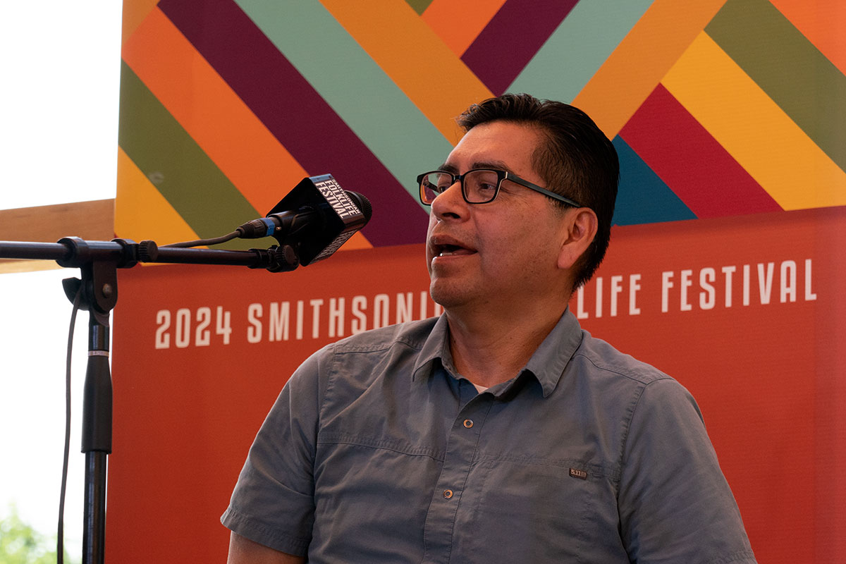 A man wearing a blue button-up shirt and black glasses speaks on an outdoor stage. The orange backdrop behind him reads 2024 Smithsonian Folklife Festival.