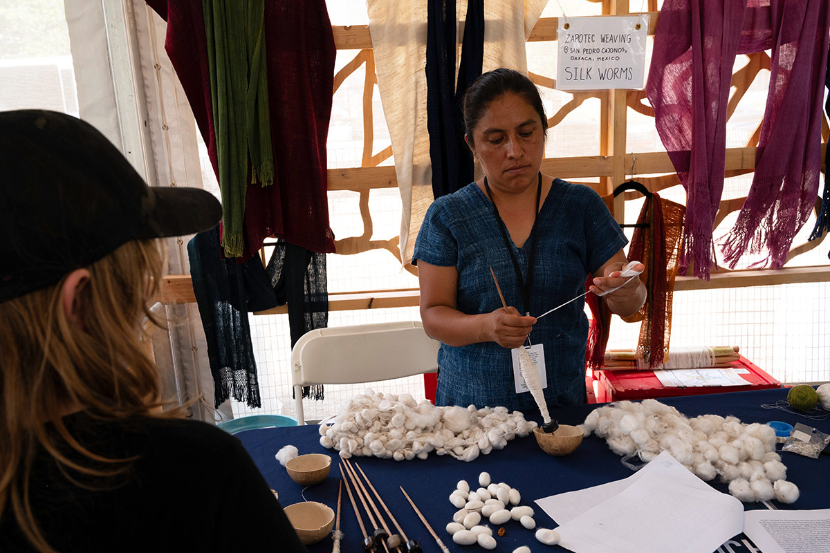 A woman twists white silk thread around a wooden stick. On the table in front of her are piles of white silkworm cocoons in various states of unraveling. Behind her, hanging on wooden trellis, are woven silk garments dyed deep red, green, and pink.