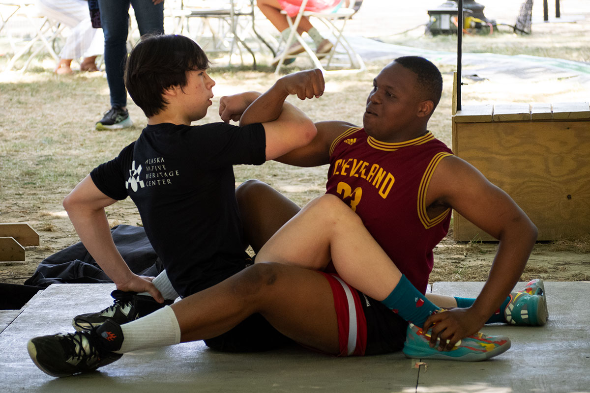 Two young men take a wrestling position seated between each other's legs on an outdoor platform, with right arms hooked at the elbows.