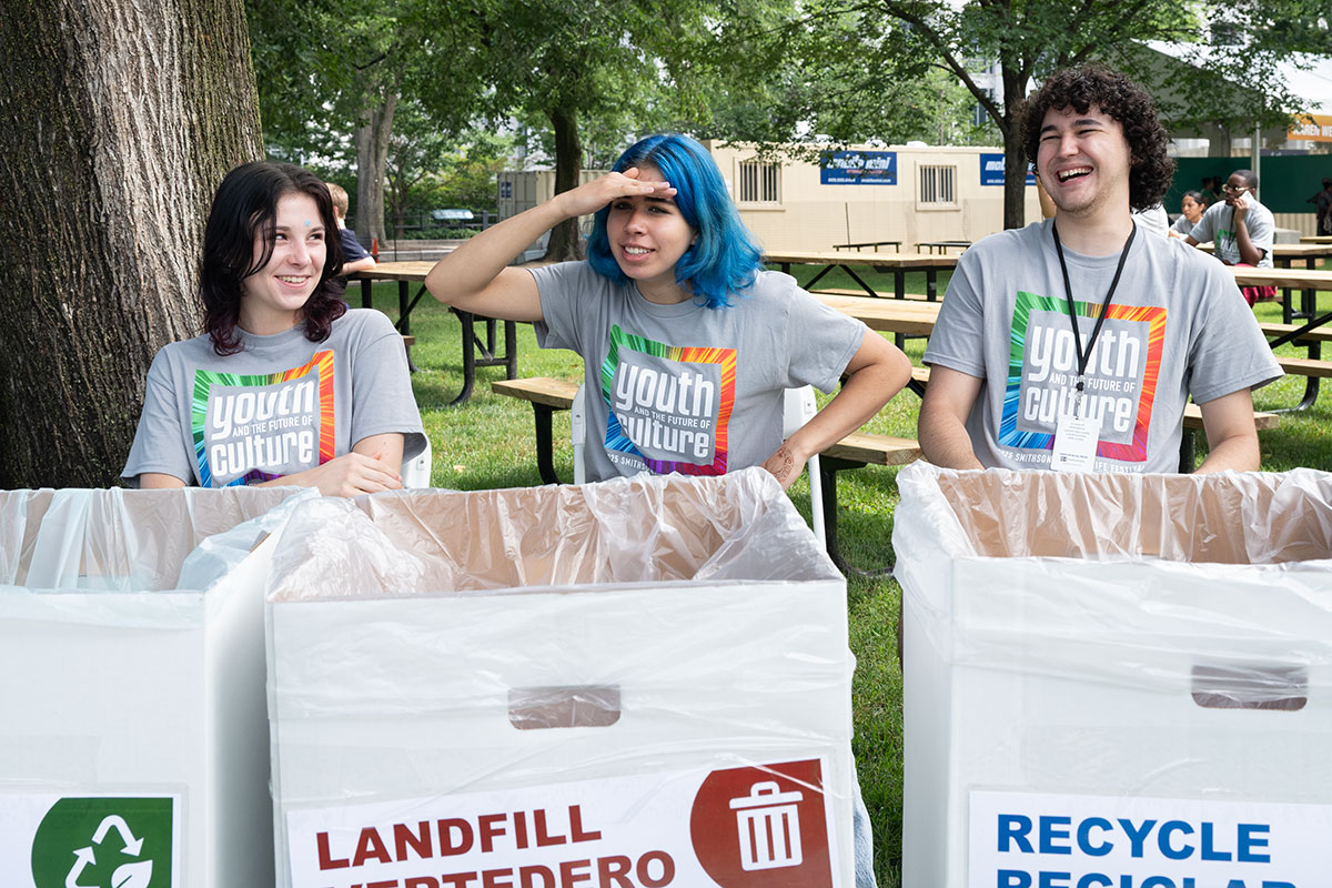 Three young people in matching gray Youth and the Future of Culture T-shirts sit behind cardboard bins marked for compose, landfill, and recycle. The person in the middle, with shoulder-length blue hair, shades her eyes looking into the distance, while the others laugh.