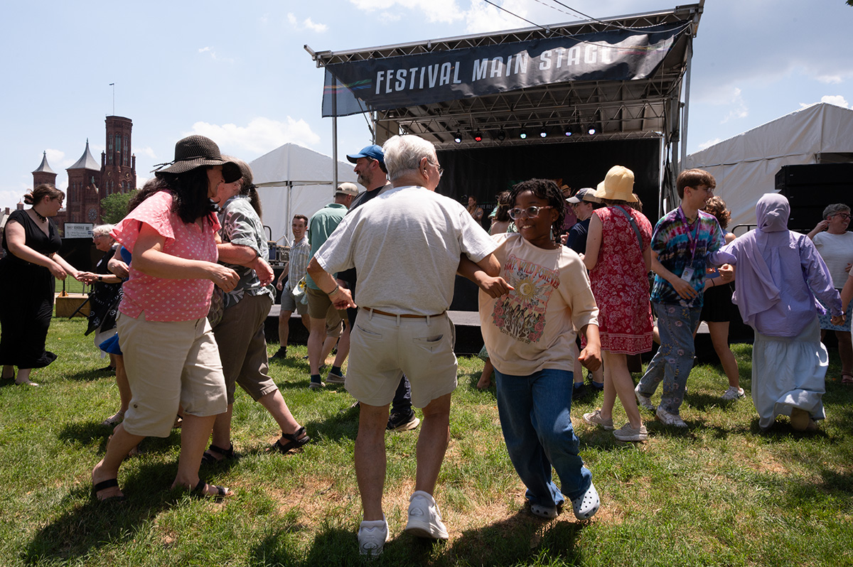 On a patch of grass in front of a stage, pairs of two people have their arms linked and spin around in circles. In the foreground, an older man and younger girl dance together.
