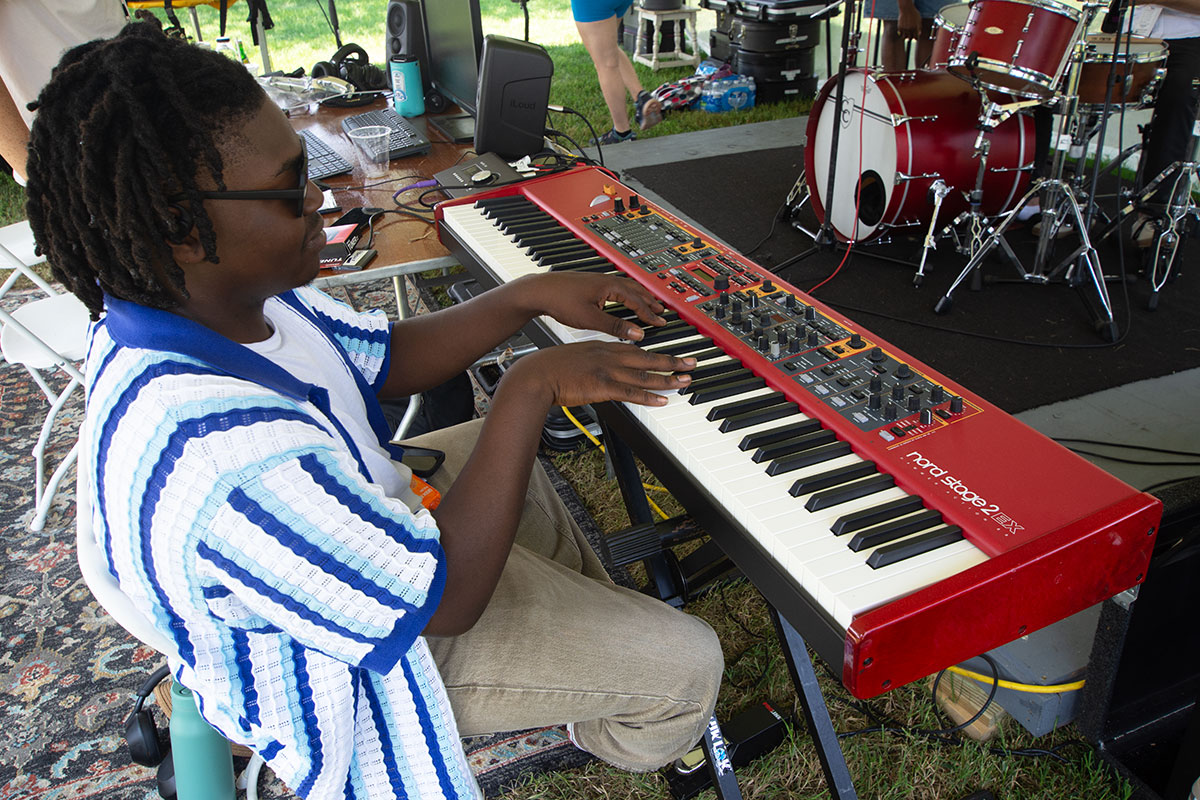 A young man wearing sunglasses and a blue, aqua, and white striped knit shirt plays a bright red keyboard set up in the grass.