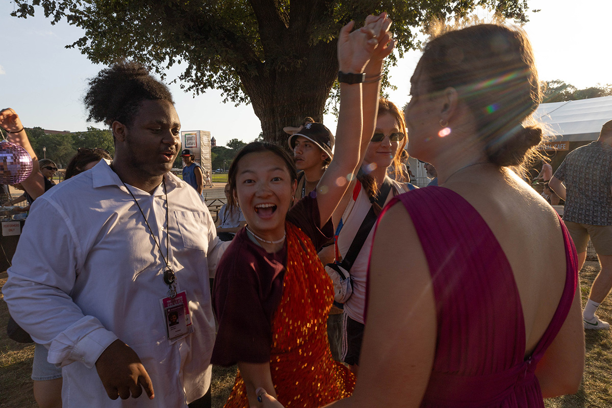A young woman wearing an orange, sparkly dress over a maroon t-shirt smiles at the camera as another young woman twirls her around. To the left, an off-camera person holds a small, purple disco ball and a young man is talking to the woman in the orange dress. To the right, another young woman is faced away from the camera wearing a wine-red dress and the sunlight creates a flare.