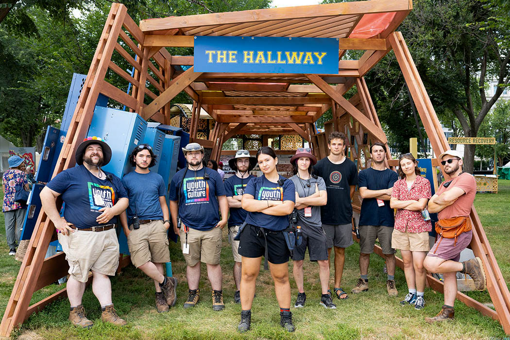 Ten members of the 2025 Smithsonian Folklife Festival's tech crew posing under the framing of The Hallway's wooden structure; six have their arms crossed, two in their pockets, and two by their sides.