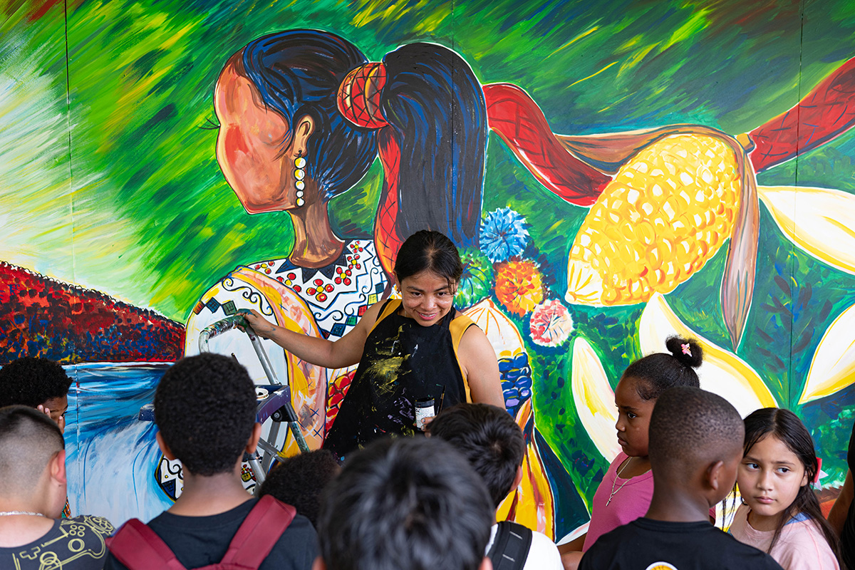 A woman with black apron covered in paint faces a group of small children, smiling and holding onto a step ladder. Behind her is a mural in vibrant colors of a woman with an embroidered blouse and long dark hair in a ponytail, plus giant ears of corn and white petals of a flowers.
