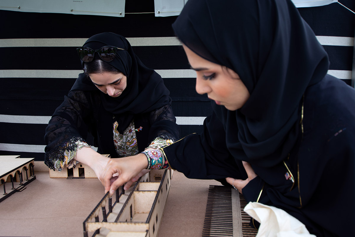 Two women lean over a small, wooden architecture model and adjust the walls. They are both looking down at the structure and in the background is a striped, black and white wall. 