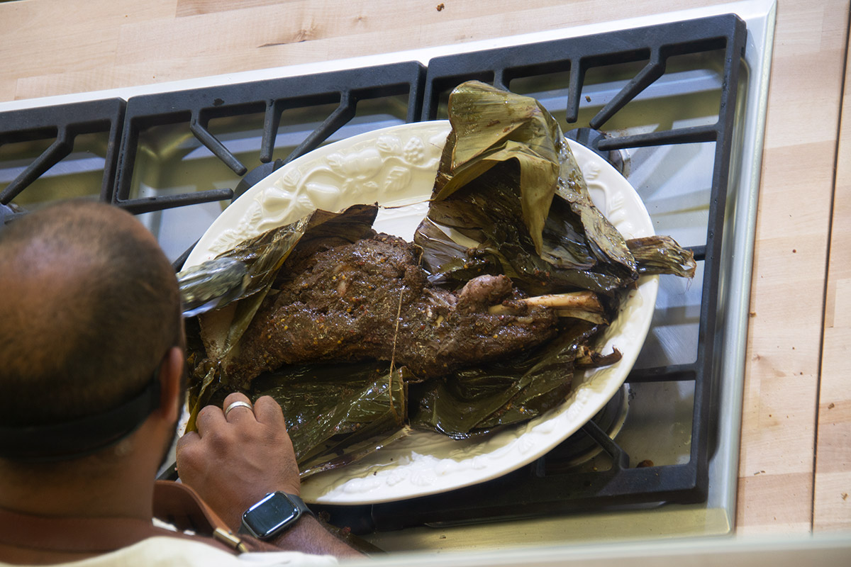 From behind, a man looks over a white oval platter of a deep brown piece of bone-in meat, unwrapping it from dark green banana leaves.