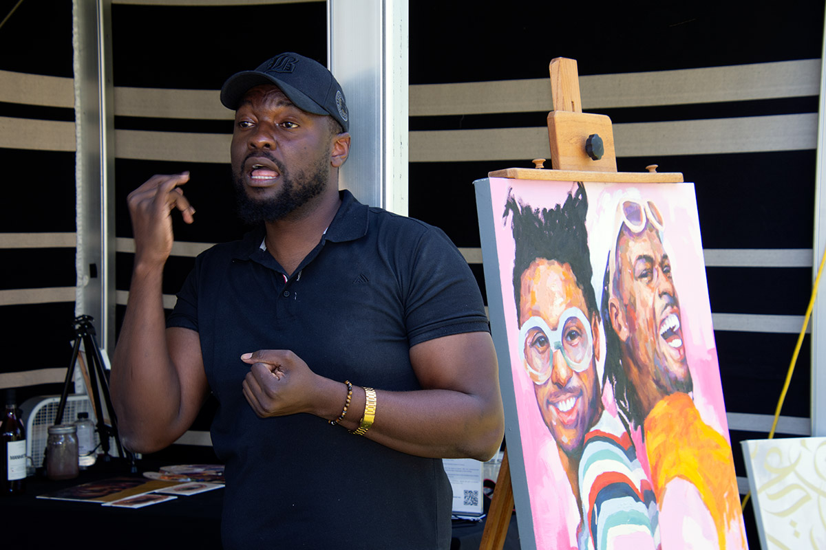 A man uses sign language, standing in front of a brightly colored painting on canvas of two men against a pink background.