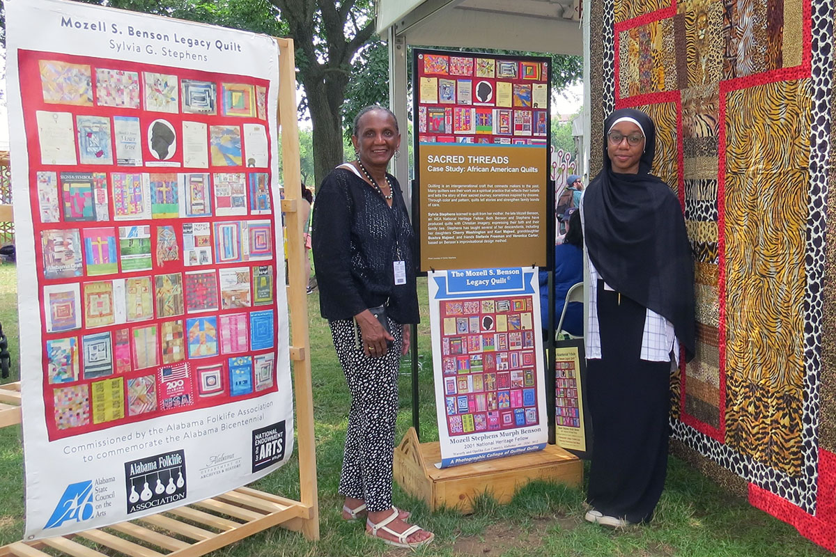 Two women stand among signs with images of quilts and a hung quilt with a Christian cross in animal prints.