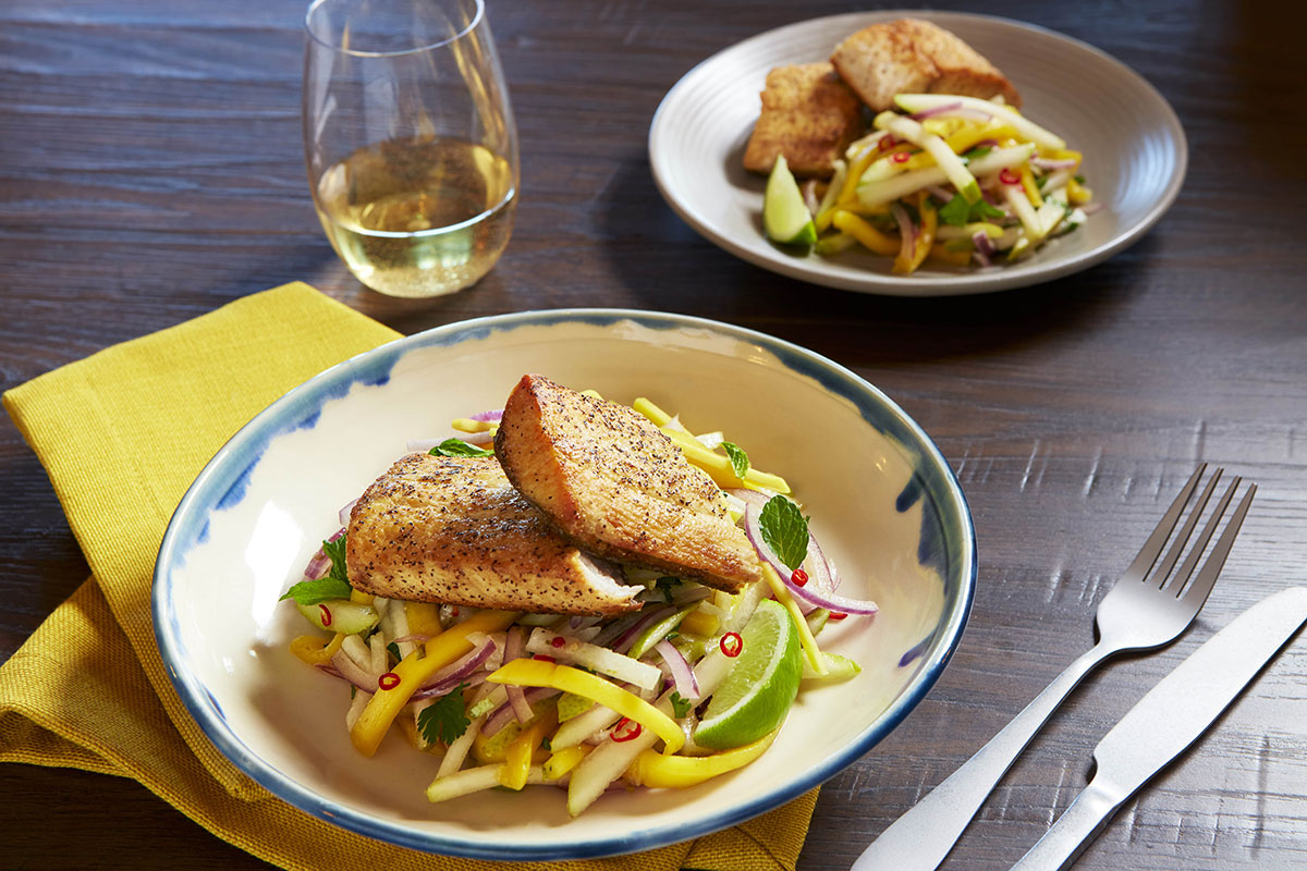 Two round dishes, each with two pieces of fish resting on a bed of chopped yellow mango, white jicama, purple onion, red peppers, green herbs, and a green lime wedge.