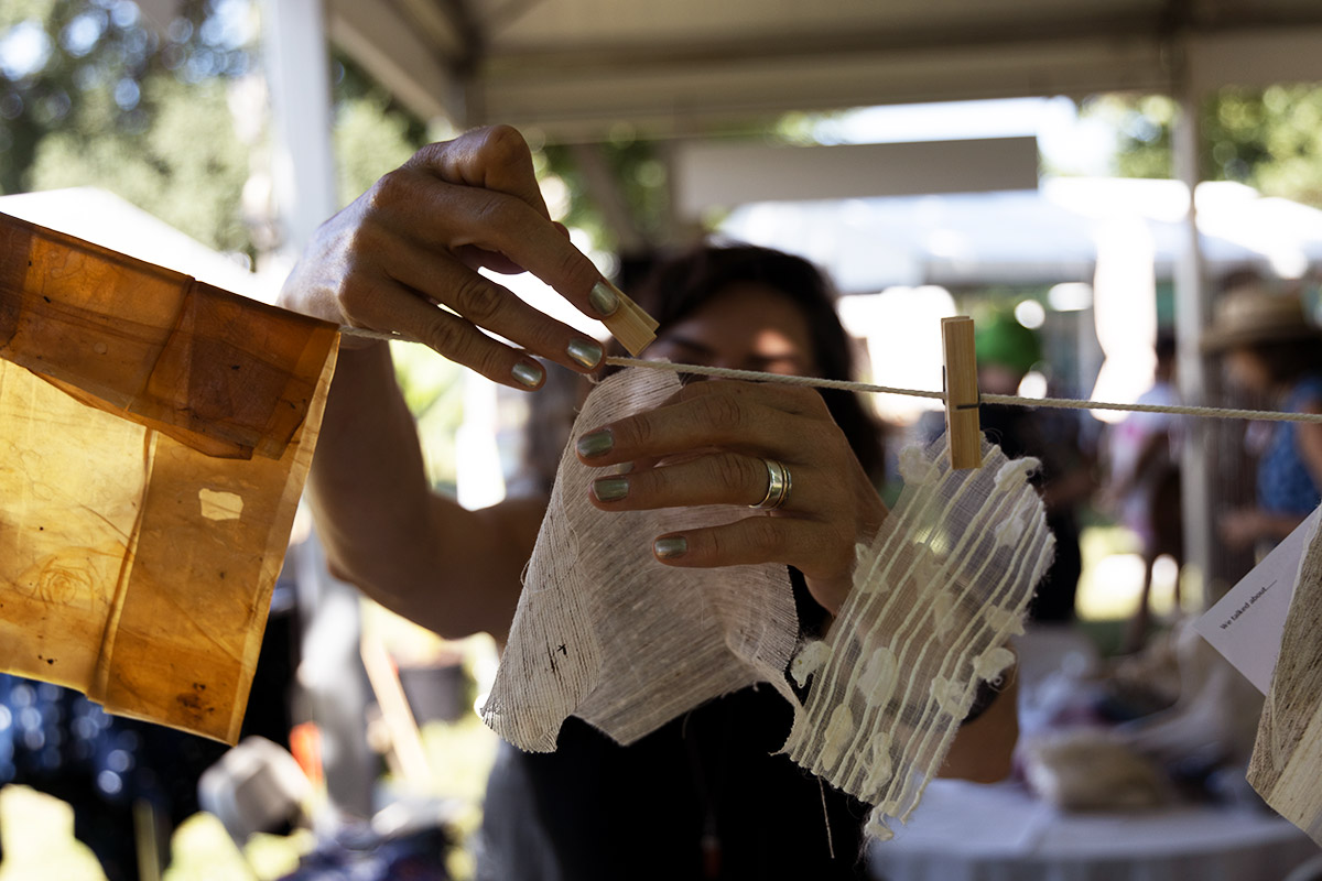 Close-up on a woman, her face obscured, as she pins a square of white fabric to a clothesline. Other fabrics, one translucent brown and the other white and lacey, are on either side.