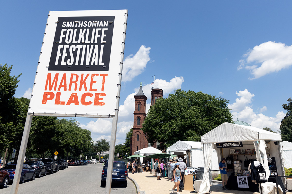 A large banner outdoors that reads Smithsonian Folklife Festival Marketplace, with white tent stalls in the background.
