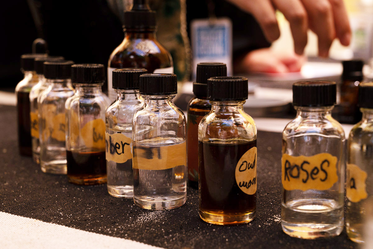 Close-up on clear class bottles filled with clear and dark brown liquids. Some are labeled: oud wood, roses, amber.