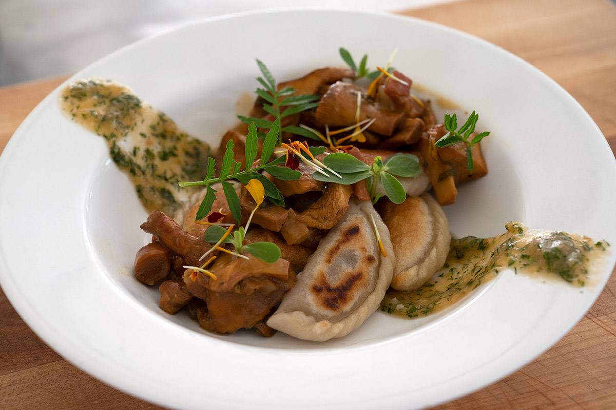 A white dish plated with tan fried dumpling, brown cooked mushrooms, a brown sauce flecked with green herbs, and garnished with sprigs of edible herbs and flowers.