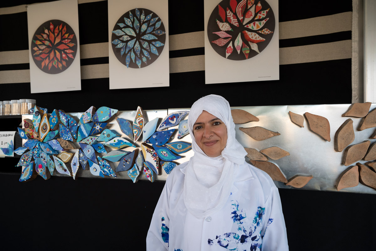 A woman in white blouse and headscarf smiles, standing in front of a silver metal surface on a wall, containing hand-painted eye-shaped magnets in shades of blue, white, and purple. Above it, photo prints of similar artworks, the magnets arranged in flower shapes.