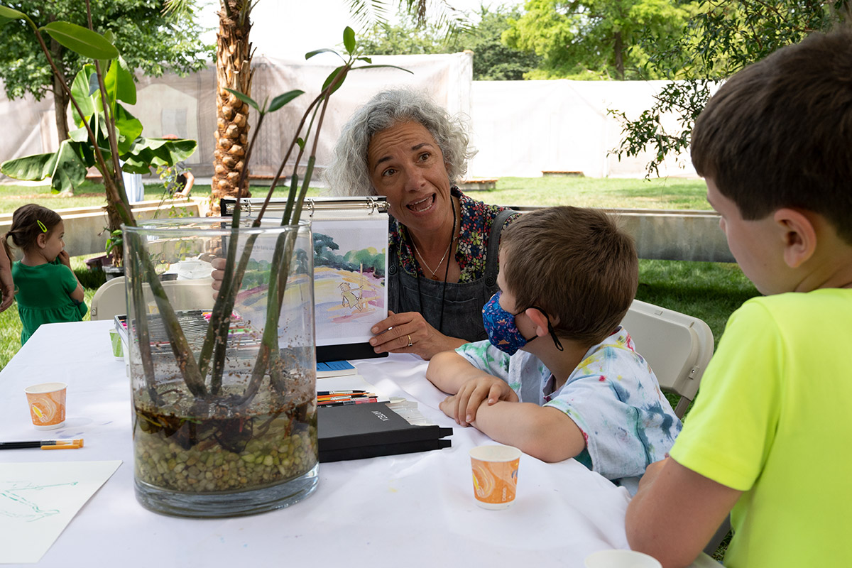A woman with short gray hair and floral shirt props up a watercolor painting to show two children sitting at a table outdoors with her. In the center of the table, a glass vase holding small mangrove stalks.