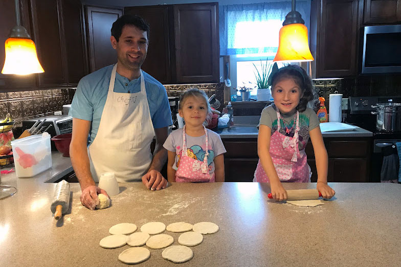 A man and two young girls pose at a kitchen counter, all wearing aprons. One girl rolls out a circle of dough, with more on the floured counter in front of them.