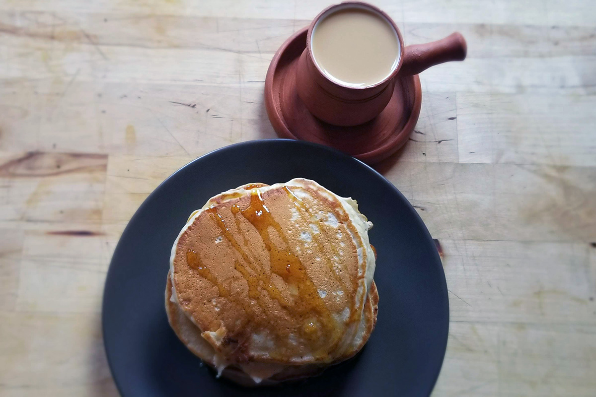 A stack of chbaab (pancakes) drizzled with syrup on a black plate, next to a red cup of milky brown chai karak.