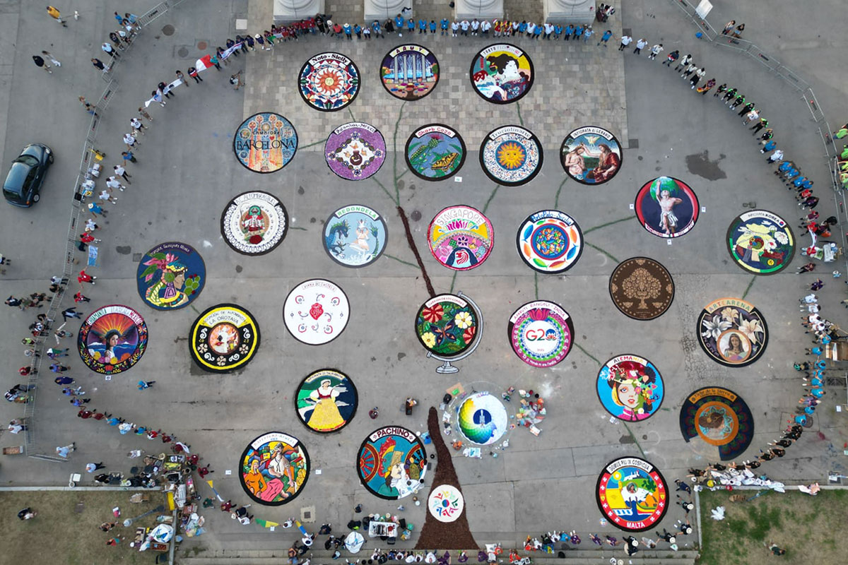 From above, several colorful, circular designs on a gray stone plaza. Lines between them, a brown trunk at the bottom, and an uneven ring of people around the perimeter suggest the shape of a tree. 