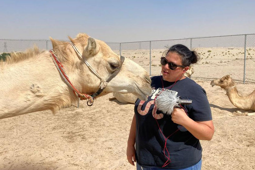 A woman holds a microphone up to the mouth of a camel. Another camel sits in the desert sand behind her.