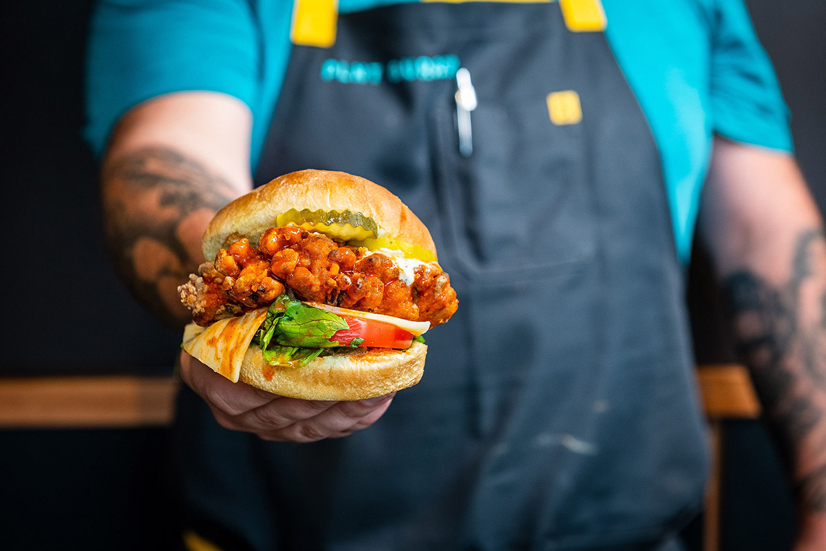 Close-up on a sandwich with breaded mushrooms, green pickles, red tomato, white cheese, and greens, held out by a person in navy apron and tattooed arms.