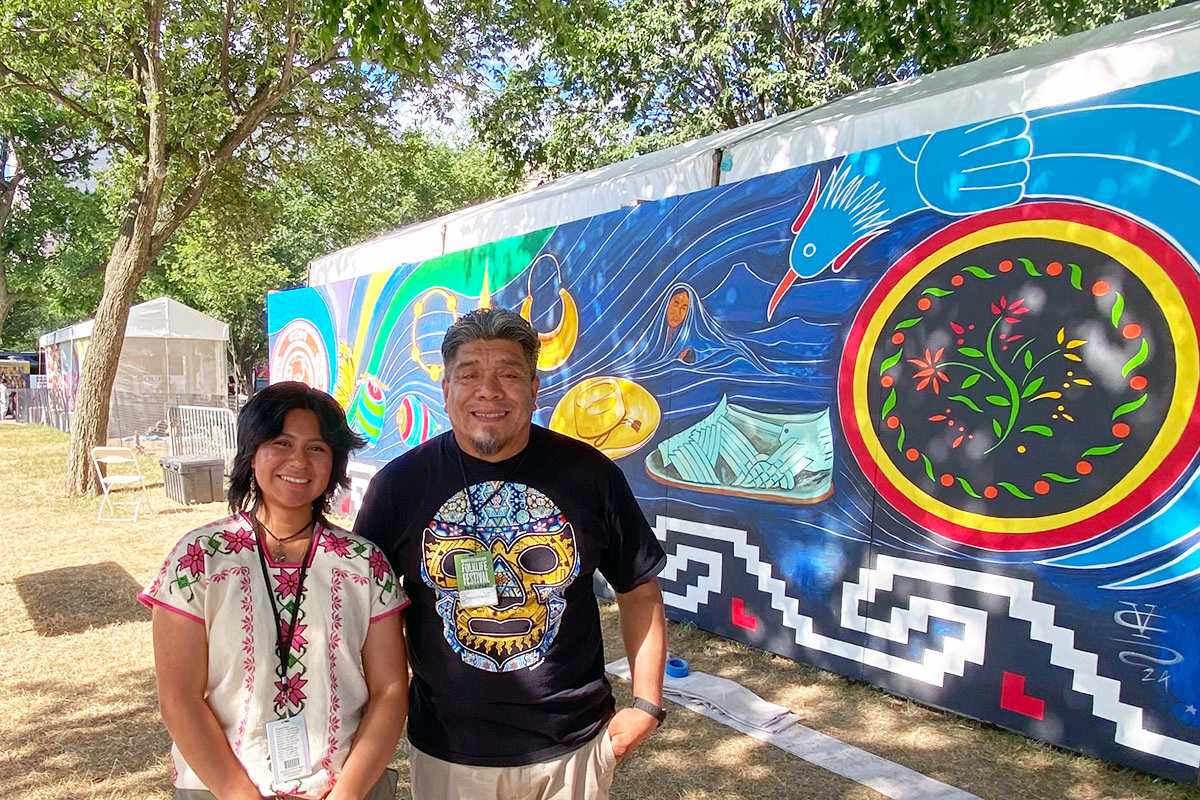 A man and a woman smile in front of an outdoor mural, depicting a leather sandal, flowers, a bird, gold jewelry, a brimmed hat, and other symbols on a dark blue background.