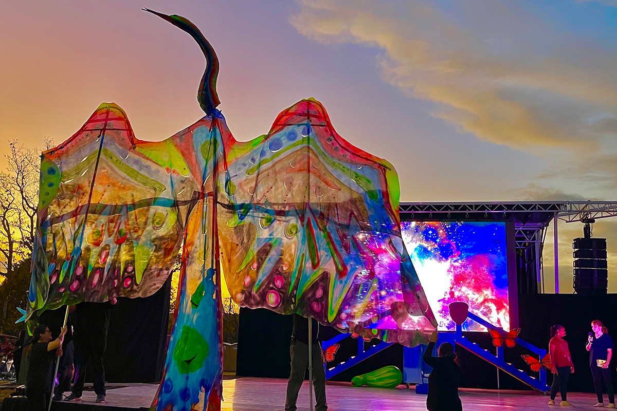 A translucent puppet of a bird with outstretched wings, each held up by a person holding a pole, takes center stage outdoors, under a pink sky at sunset. 