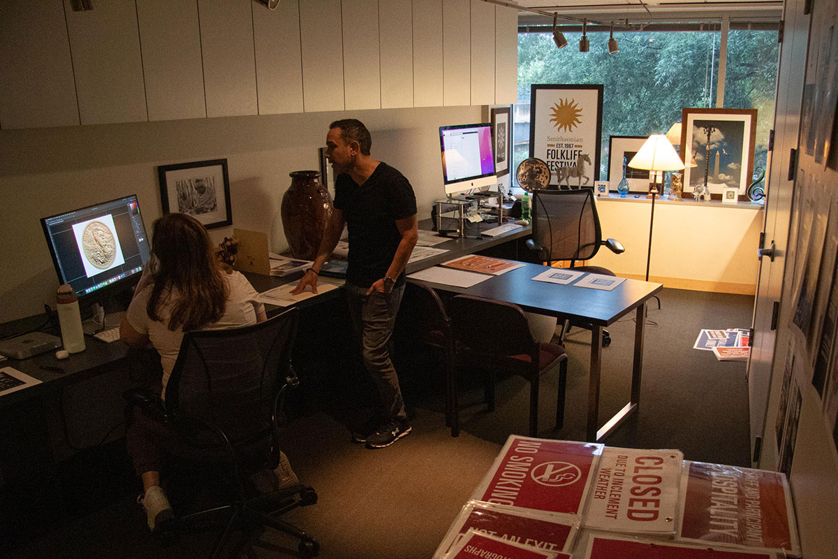 In a warmly lit office room, a young woman works at a computer, consulting with a man standing beside her. Taped to the walls and covering the tabletops are laminated signs and printouts of designs.