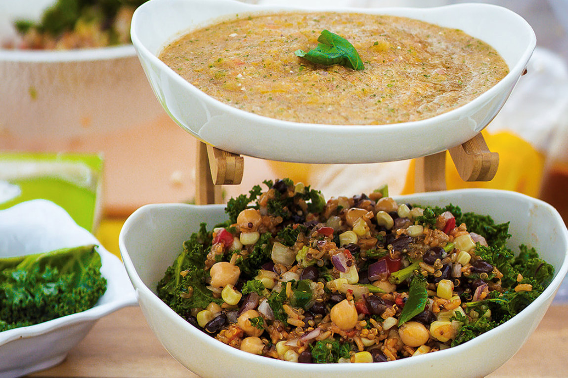 A dish of yellow tomato soup with a green basil leaf on top and a dish of salad with green kale leaves, red beans, purple onion, chickpeas, and chopped red pepper.