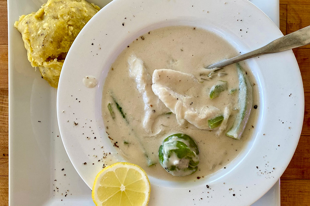 A white bowl filled with creamy white soup with chunk of white fish and green peppers and okra, and a yellow lemon slice on the rim. On a square plate below it, a mound of yellow plantain mash.