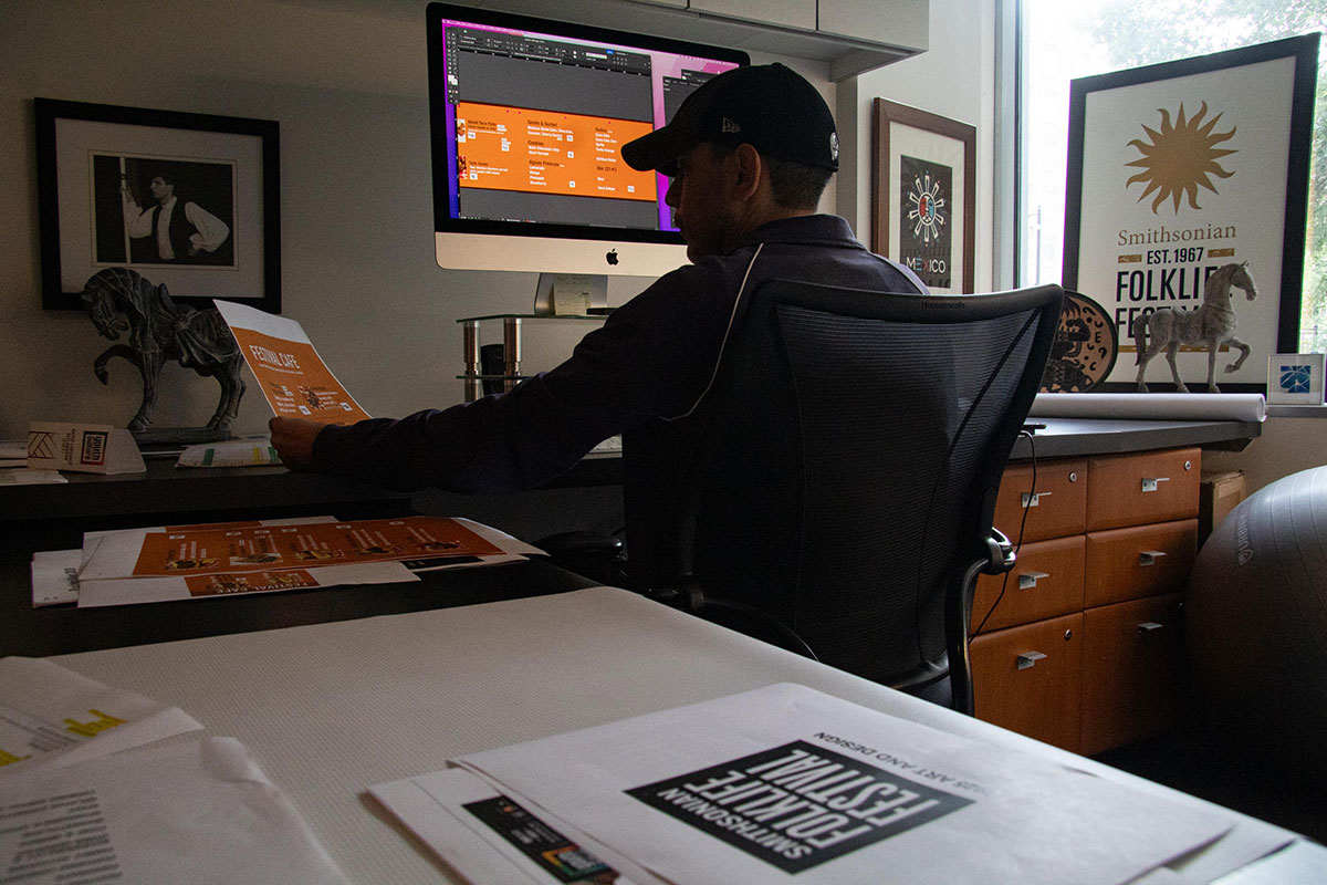 A man sits in a dimly lit room, working on a computer with printouts of Folklife Festival signage on the desks around him.