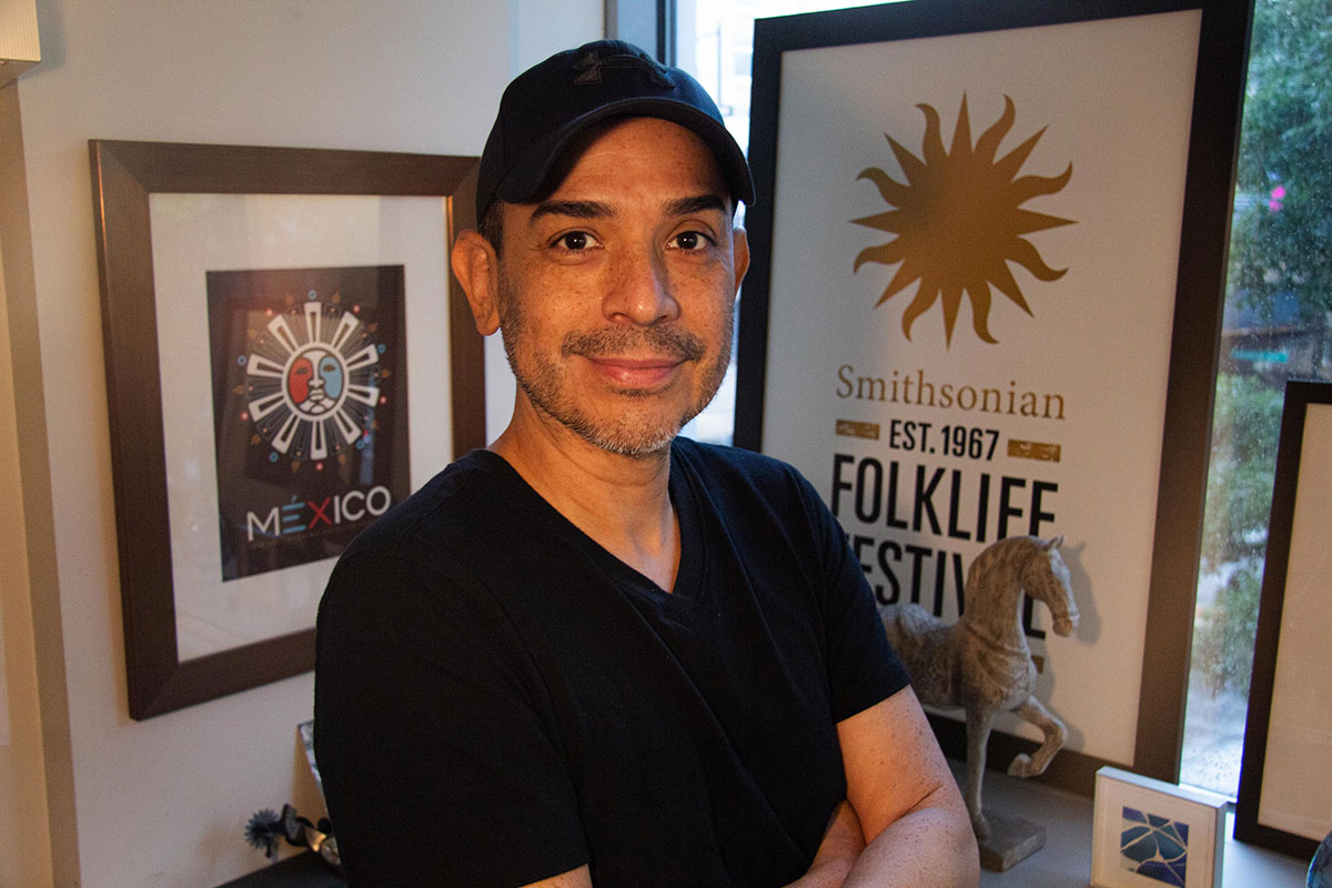 A man wearing black ballcap and V-neck shirt smiles at the camera in warm lighting. Behind him, framed on the walls, are logos for the Smithsonian Folklife Festival and the Mexico program, both featuring sun imagery.