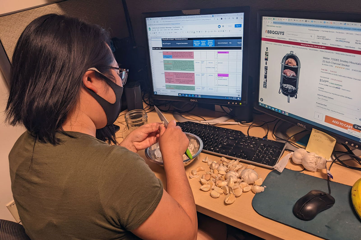 Facing away, a woman sits at a desk, peeling several cloves of garlic in front of a computer.