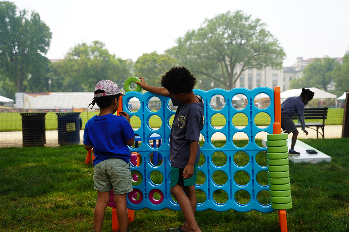 Two children face away from the camera while playing on a large Connect Four set in some grass outdoors.