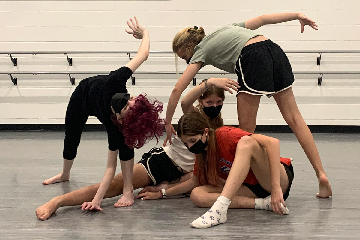 Four young dancers on a gray studio floor, arms and legs outstretched in curving shapes.
