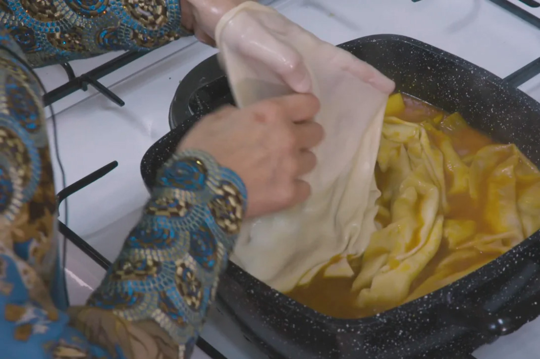 Closeup on a pair of hands layering thin sheets of dough or noodle in a square pot with red broth on a stovetop.